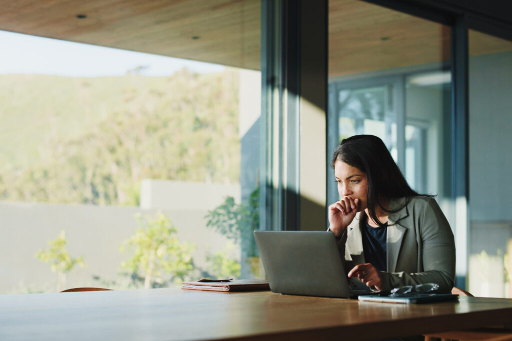 Woman in a blazer works on a laptop at a wooden table by large glass windows with greenery outside.