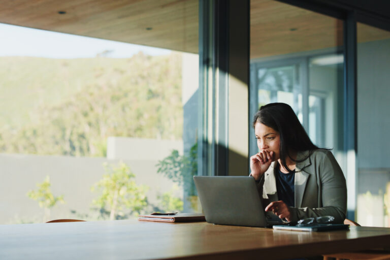 Woman in a blazer works on a laptop at a wooden table by large glass windows with greenery outside.