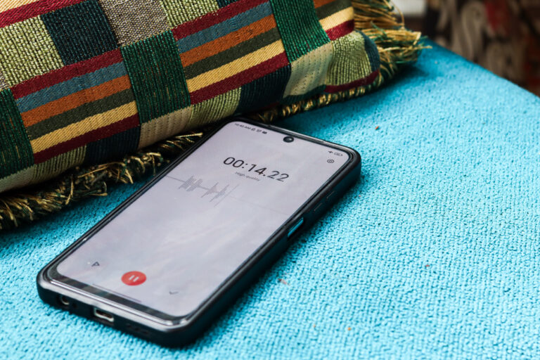 Smartphone lying on a blue towel near a colorful patterned cushion, screen showing a recording app with a red pause/record button.