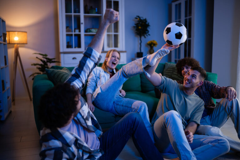 Group of excited friends celebrating goal with a soccer ball while watching sports tv at home, enjoying together